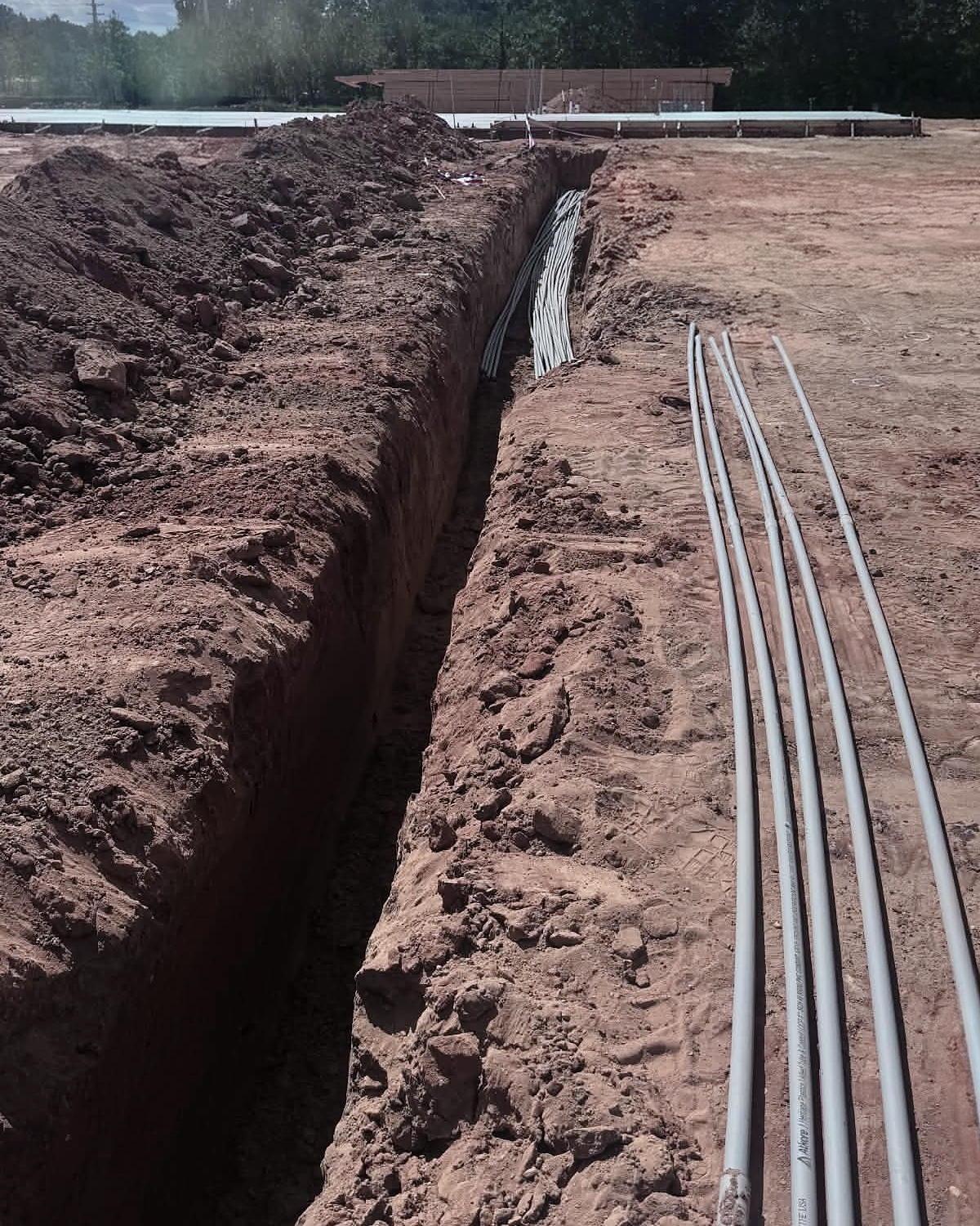 Open trench cut through red clay, four conduit runs laid beside it, the building slab in the distance