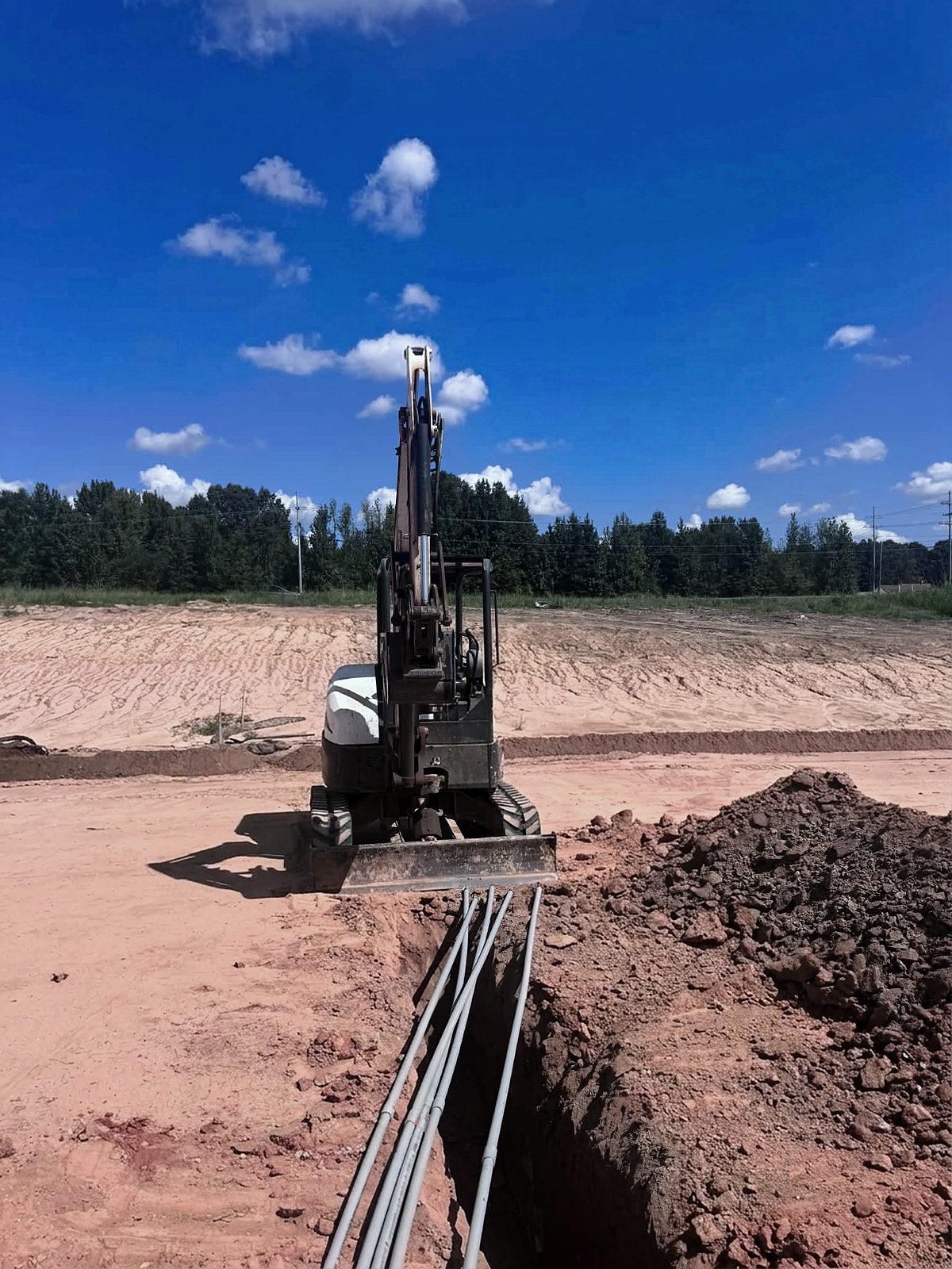 Mini excavator parked over an open trench, four conduit runs entering it from the foreground