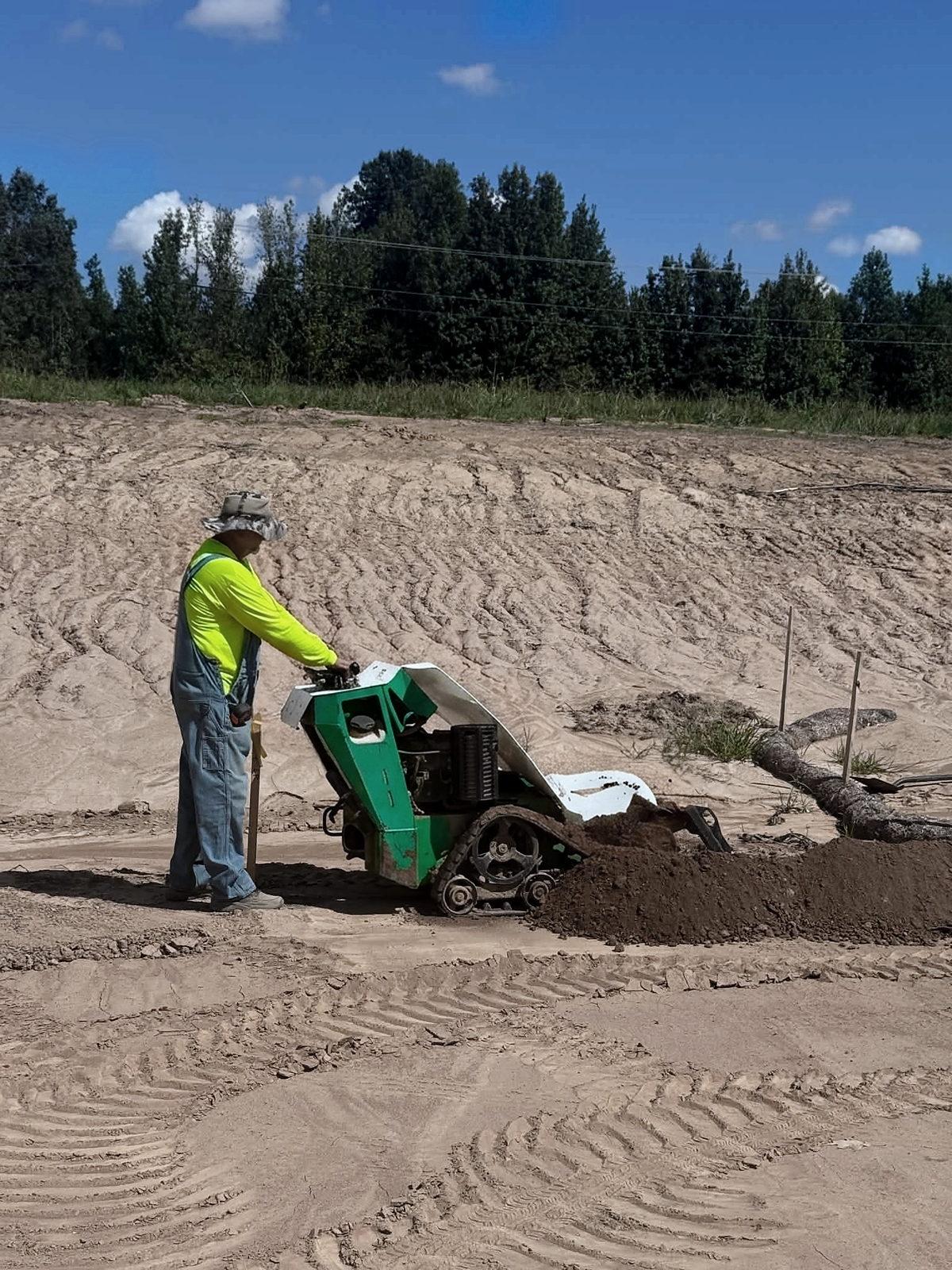 An electrician guiding a walk-behind trencher along the line, spoil piling beside the cut