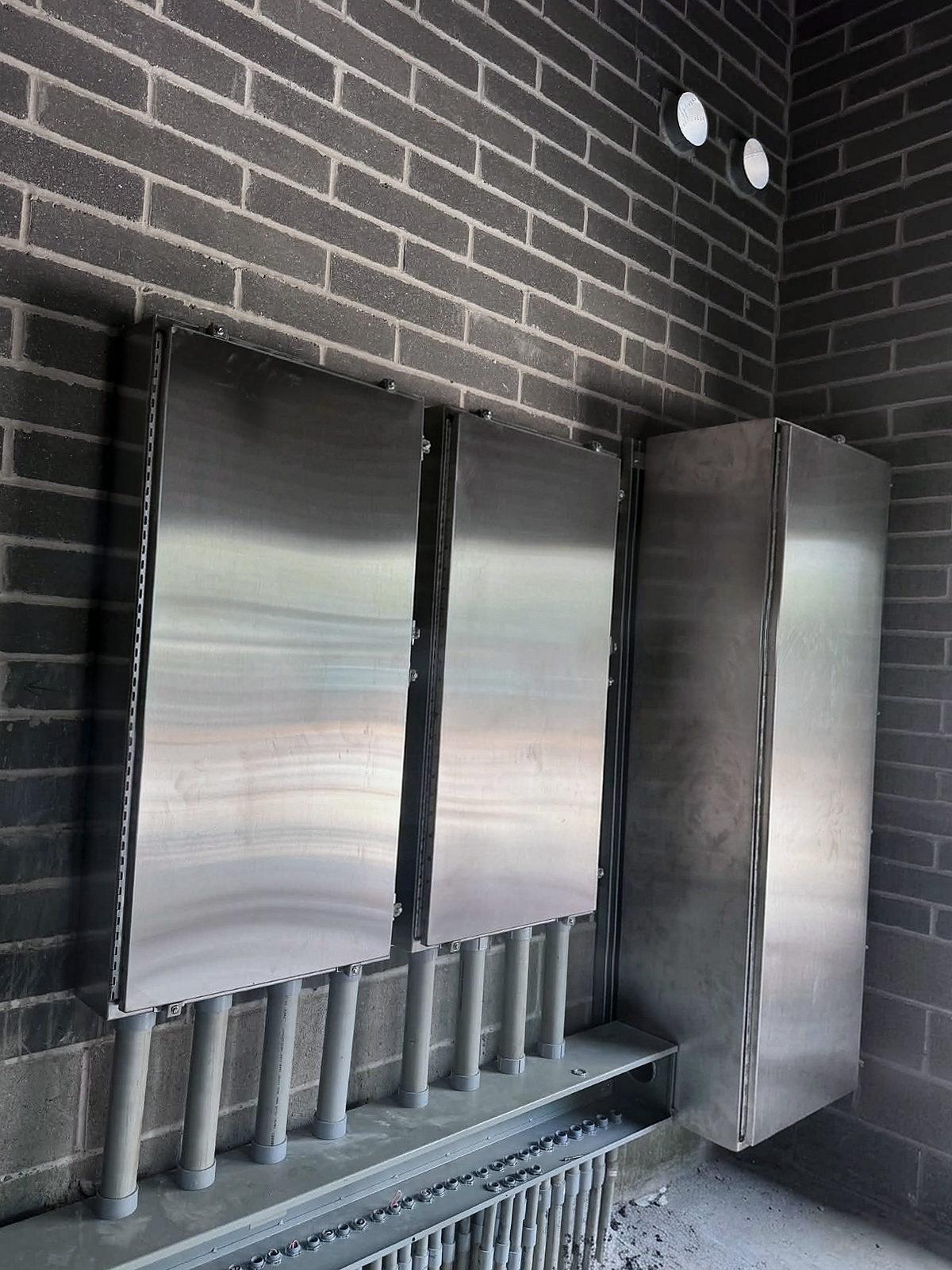 Three stainless steel enclosures mounted on a brick wall, conduit stubs running up into them from below