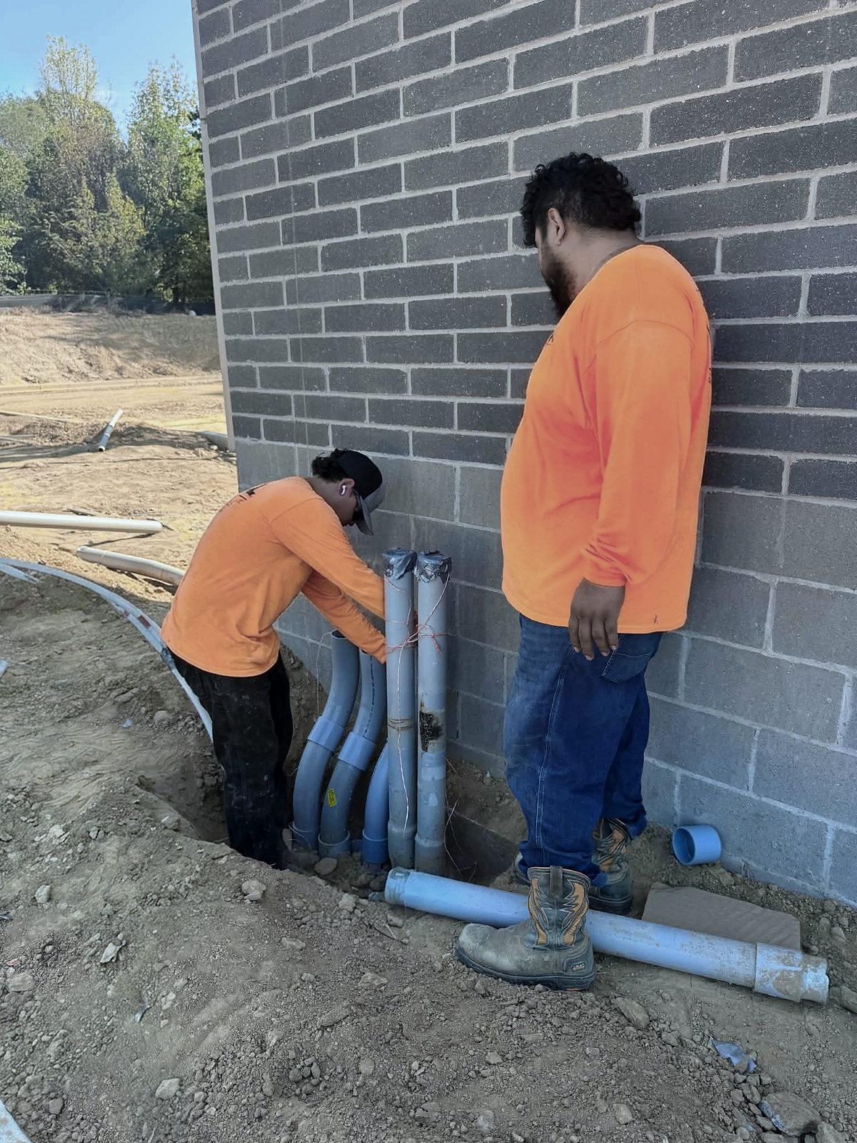 Two electricians setting conduit risers against a block wall, the bends stubbed up from the footing